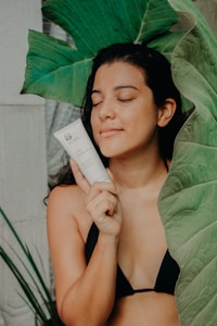 A person in a black bikini holds a skincare product near their face, surrounded by large green leaves. Their eyes are closed, and they appear relaxed and content.