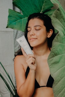 A person in a black bikini holds a skincare product near their face, surrounded by large green leaves. Their eyes are closed, and they appear relaxed and content.