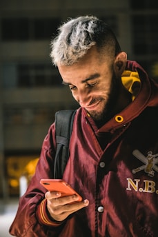 A man with a short beard and styled hair is wearing a maroon jacket with a baseball logo. He is holding and looking at a smartphone, appearing focused and slightly amused. The background is blurred, highlighting the man as the focal point of the image.