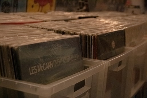 Stacks of vinyl records organized in plastic storage bins, with visible album covers, including one labeled 'Les McCann'. The scene is dimly lit, suggesting a cozy or vintage atmosphere.