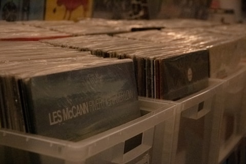 Stacks of vinyl records organized in plastic storage bins, with visible album covers, including one labeled 'Les McCann'. The scene is dimly lit, suggesting a cozy or vintage atmosphere.