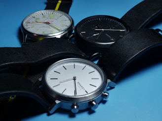 Three wristwatches with round faces and black leather straps are positioned on a light blue surface. The watches have different face colors: one with a white face and green numerals, another with a black face, and the last with a silver face. The watches have various brand markings and display different times.