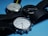 Three wristwatches with round faces and black leather straps are positioned on a light blue surface. The watches have different face colors: one with a white face and green numerals, another with a black face, and the last with a silver face. The watches have various brand markings and display different times.