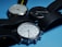 Three wristwatches with round faces and black leather straps are positioned on a light blue surface. The watches have different face colors: one with a white face and green numerals, another with a black face, and the last with a silver face. The watches have various brand markings and display different times.