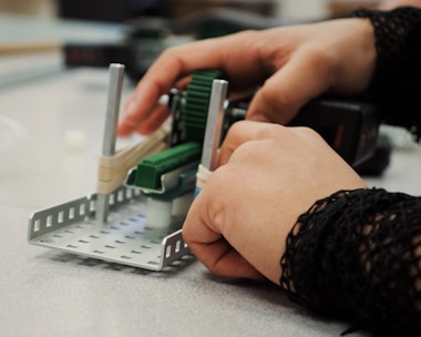 Hands are manipulating a small mechanical assembly, which includes metal and plastic components. The setup seems to involve green gears and metallic rods, indicative of a robotics or engineering-related activity.