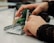 Hands are manipulating a small mechanical assembly, which includes metal and plastic components. The setup seems to involve green gears and metallic rods, indicative of a robotics or engineering-related activity.