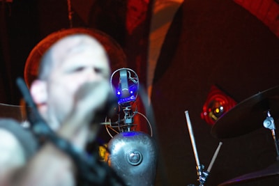 A performer is seen on stage, slightly out of focus, with a microphone in front of them. In the foreground, there is a robot-like structure with illuminated blue lights. The background has a dark, atmospheric setting with red and orange lighting accentuating the scene.