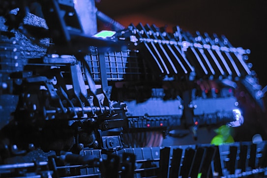 Close-up view of intricate machinery with a complex array of bolts, levers, and metal components, illuminated by a dramatic blue light. The shadows and lighting create a stark contrast, emphasizing the industrial engineering and detail.