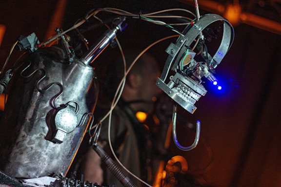 A metallic, robotic sculpture with visible wires and components is prominently featured, with a mechanical arm or appendage extending outward. The environment is dimly lit, and the focus is on the robot's intricate design and construction.