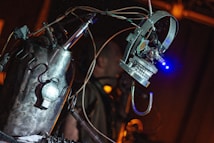 A metallic, robotic sculpture with visible wires and components is prominently featured, with a mechanical arm or appendage extending outward. The environment is dimly lit, and the focus is on the robot's intricate design and construction.