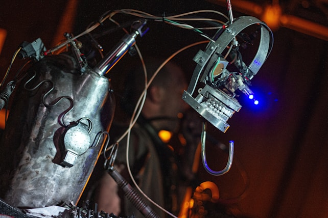 A metallic, robotic sculpture with visible wires and components is prominently featured, with a mechanical arm or appendage extending outward. The environment is dimly lit, and the focus is on the robot's intricate design and construction.