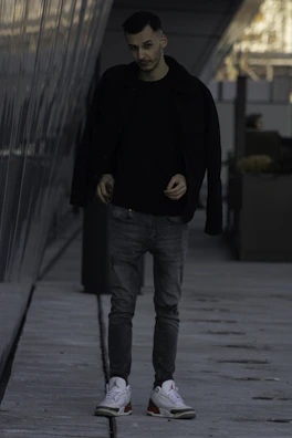 Model walking through an urban alley wearing layered black streetwear with subtle reflective accents.