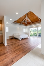 A spacious and modern bedroom features a wooden floor and a large bed with white bedding. The room is illuminated by natural light coming through a sliding glass door, which opens up to a lush, green outdoor view. The ceiling has a wooden finish with a traditional fan in the center, adding a touch of rustic charm.