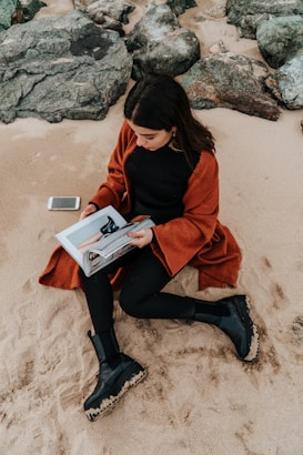 A woman is sitting on a sandy beach, wearing a rust-colored coat and black boots. She is holding a magazine open in her hands, and there is a smartphone placed beside her on the sand. The beach is bordered by large gray rocks.