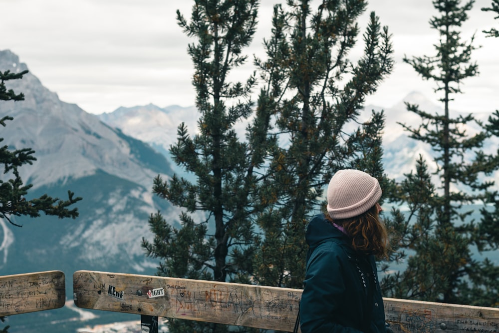 woman in blue jacket and white knit cap standing near brown wooden fence looking at snow