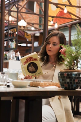 A woman sitting in a cafe on the phone while reading a book titled 'Cora Coralina'. The setting has a warm and cozy atmosphere with visible exposed brick walls and hanging lights. There are plants and a mug on the table along with other dining items.