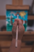 A child's hand is holding a paintbrush against a colorful canvas on an easel. The painting features abstract shapes and bright colors, including a sun-like figure. The focus is on the child's hand, emphasizing the act of painting.