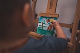 A child is painting on a small canvas positioned on an easel. The painting depicts abstract shapes and colors, suggesting plants and a sunny sky with blue, green, and brown colors. The child is holding a brush and is focused on adding details to the artwork.