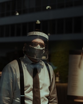 A person wearing a face shield with the word 'ZUCCA' on it, along with a face mask, a white shirt, and suspenders. The background appears to be a dimly lit urban environment.