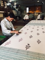 A craftsman is engaged in block printing on a large piece of fabric. He works meticulously with wooden stamps to create intricate floral patterns. The workshop has a traditional and rustic ambiance, with other workers in the background focused on similar tasks. The workspace is filled with various wooden tools and completed textiles, and it is lit with overhead lights.