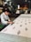 A craftsman is engaged in block printing on a large piece of fabric. He works meticulously with wooden stamps to create intricate floral patterns. The workshop has a traditional and rustic ambiance, with other workers in the background focused on similar tasks. The workspace is filled with various wooden tools and completed textiles, and it is lit with overhead lights.