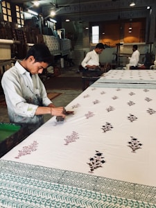 A craftsman is engaged in block printing on a large piece of fabric. He works meticulously with wooden stamps to create intricate floral patterns. The workshop has a traditional and rustic ambiance, with other workers in the background focused on similar tasks. The workspace is filled with various wooden tools and completed textiles, and it is lit with overhead lights.