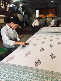 A craftsman is engaged in block printing on a large piece of fabric. He works meticulously with wooden stamps to create intricate floral patterns. The workshop has a traditional and rustic ambiance, with other workers in the background focused on similar tasks. The workspace is filled with various wooden tools and completed textiles, and it is lit with overhead lights.