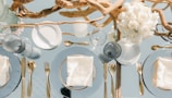An elegantly set dining table features gold cutlery and glass plates with neatly folded white napkins. A centerpiece composed of intertwined driftwood and a bouquet of white roses adds a natural and sophisticated touch. The table setting is complemented by clear glassware that reflects light, enhancing the luxurious ambiance.