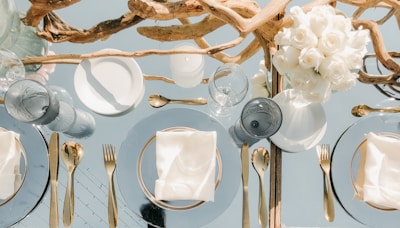 An elegantly set dining table features gold cutlery and glass plates with neatly folded white napkins. A centerpiece composed of intertwined driftwood and a bouquet of white roses adds a natural and sophisticated touch. The table setting is complemented by clear glassware that reflects light, enhancing the luxurious ambiance.