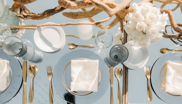 An elegantly set dining table features gold cutlery and glass plates with neatly folded white napkins. A centerpiece composed of intertwined driftwood and a bouquet of white roses adds a natural and sophisticated touch. The table setting is complemented by clear glassware that reflects light, enhancing the luxurious ambiance.