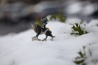 A small figurine of a mythical creature is positioned on a snowy landscape surrounded by patches of green leaves. The creature has a dark body with a distinctive headdress and a wolf-like appearance. The focus is on the figurine, with the background softly blurred, suggesting an outdoor setting.