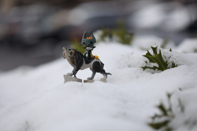 A small figurine of a mythical creature is positioned on a snowy landscape surrounded by patches of green leaves. The creature has a dark body with a distinctive headdress and a wolf-like appearance. The focus is on the figurine, with the background softly blurred, suggesting an outdoor setting.