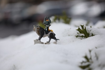 A small figurine of a mythical creature is positioned on a snowy landscape surrounded by patches of green leaves. The creature has a dark body with a distinctive headdress and a wolf-like appearance. The focus is on the figurine, with the background softly blurred, suggesting an outdoor setting.