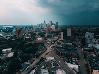 A cityscape depicting an urban area with multiple high-rise buildings under a cloudy sky. The infrastructure includes roads, intersections, and a mix of taller office blocks and smaller structures. The horizon is filled with a dramatic transition from a bright to a dark, stormy sky.