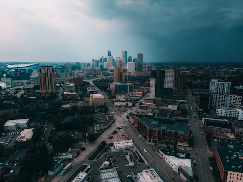 A cityscape depicting an urban area with multiple high-rise buildings under a cloudy sky. The infrastructure includes roads, intersections, and a mix of taller office blocks and smaller structures. The horizon is filled with a dramatic transition from a bright to a dark, stormy sky.