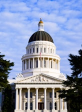 A large, classical-style building with a prominent white dome and detailed architectural features stands under a bright sky. The structure is adorned with columns and decorative sculptures at the front. An American flag is visible at the top of the building.