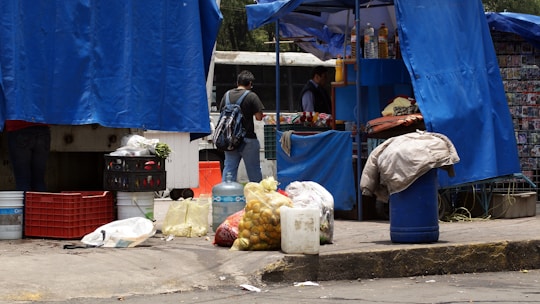 A street market stall is partially covered with vibrant blue tarps. Various goods, including bags of produce and large bottles of water, are stacked on the ground. A person with a backpack stands nearby, possibly browsing or purchasing items from the vendor inside the stall.