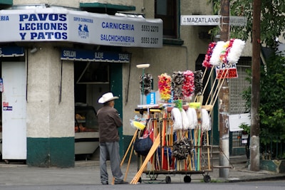 A street scene featuring a man wearing a white hat and grey pants standing next to a cart filled with various cleaning tools such as brooms, mops, and feather dusters. Behind him is a building with signs in Spanish advertising a butcher shop that sells turkeys, piglets, and ducks. The street sign reads 'Mazatlan.'