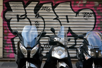 Three motorcycles are parked in front of a metal garage door with colorful graffiti featuring bold black and pink patterns. The motorcycles have clear windshields and are positioned closely in a line.