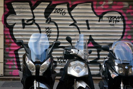 Three motorcycles are parked in front of a metal garage door with colorful graffiti featuring bold black and pink patterns. The motorcycles have clear windshields and are positioned closely in a line.