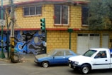 A yellow brick building with red trim and barred windows stands at a street corner. The garage door features a mural with abstract blue and black designs incorporating circular shapes. A green traffic light is positioned on the corner. Two vehicles, a blue sedan and a white pickup truck, are parked nearby. The truck has a person inside.