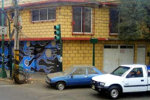 A yellow brick building with red trim and barred windows stands at a street corner. The garage door features a mural with abstract blue and black designs incorporating circular shapes. A green traffic light is positioned on the corner. Two vehicles, a blue sedan and a white pickup truck, are parked nearby. The truck has a person inside.