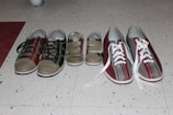 Several pairs of shoes placed on a speckled tile floor. The shoes include a pair of dark sneakers with multi-colored stitching, a pair of children's shoes with Velcro straps, and a pair of maroon and gray bowling shoes with white laces.