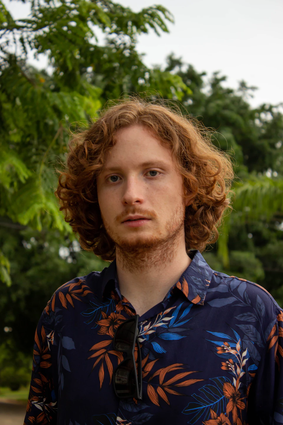 In the image, a young man with curly hair is standing against a backdrop of lush green trees and foliage. He's wearing a blue floral shirt that stands out against his vibrant red hair. The man has a serious expression on his face, giving off an air of contemplation or deep thought. His sunglasses are perched atop his head, adding to the overall cool and collected vibe he exudes.