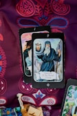 A display of tarot cards on a richly patterned fabric. The card in the foreground is The Hermit, showing an elderly figure holding a lantern and a staff, dressed in a hooded cloak. Ornate designs in red and blue create an intricate background. A woven pouch and wooden blocks are partially visible at the bottom.