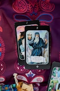 A display of tarot cards on a richly patterned fabric. The card in the foreground is The Hermit, showing an elderly figure holding a lantern and a staff, dressed in a hooded cloak. Ornate designs in red and blue create an intricate background. A woven pouch and wooden blocks are partially visible at the bottom.