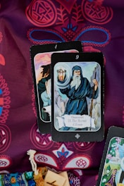 A display of tarot cards on a richly patterned fabric. The card in the foreground is The Hermit, showing an elderly figure holding a lantern and a staff, dressed in a hooded cloak. Ornate designs in red and blue create an intricate background. A woven pouch and wooden blocks are partially visible at the bottom.