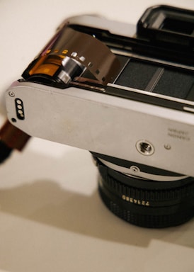A vintage camera with a roll of undeveloped film extending from the film compartment. The film is partially wound out, showcasing its dark color and perforations. The metallic body of the camera reflects light subtly, highlighting its sleek design. The lens is visible at the bottom, with engraved numbers indicating focus or aperture settings.