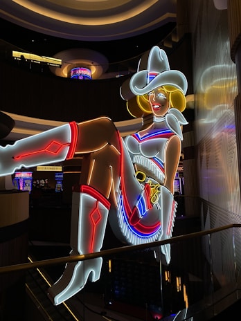 A brightly lit neon sign of a cowgirl wearing a white hat, boots, and a red, white, and blue outfit. The figure is sitting with one leg raised and crossed, in a lively and colorful style, suggestive of a vintage or retro aesthetic.