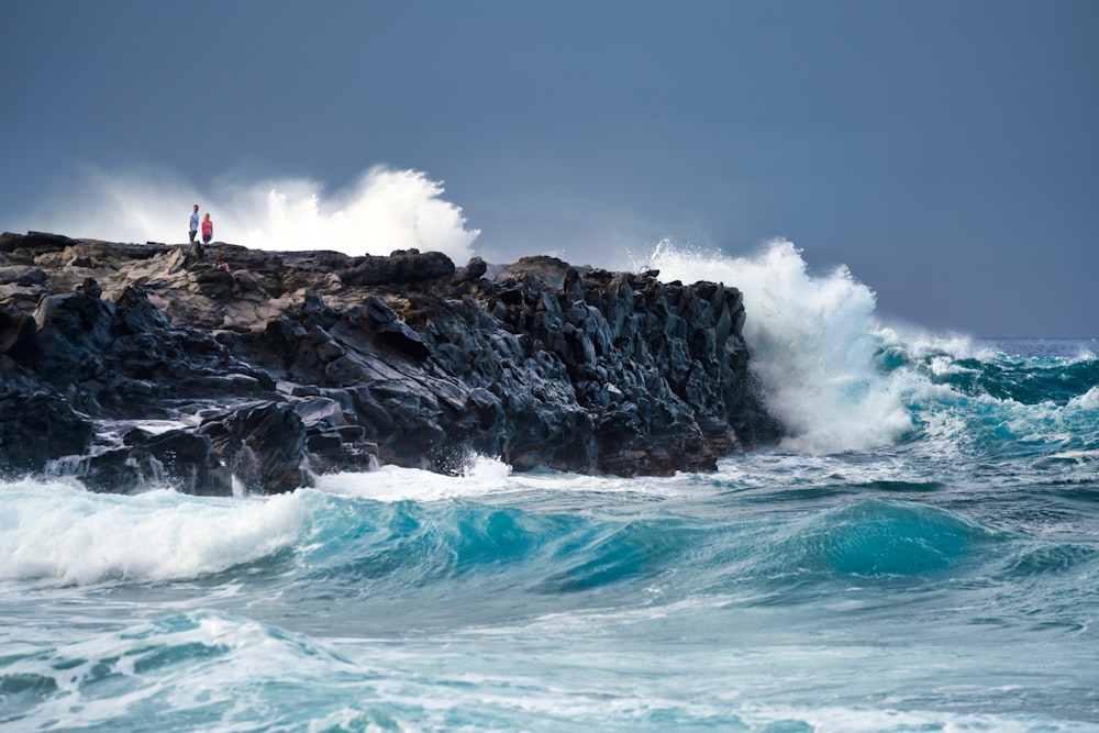 pessoa em camisa vermelha em pé na formação rochosa perto das ondas do mar durante o dia
